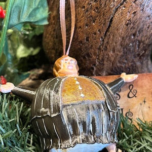 Puede incluir: Un adorno navide&ntilde;o con forma de personaje con cuerpo redondo y brazos extendidos. El adorno tiene un traje a rayas blancas y negras, un pecho dorado y una cabeza color melocot&oacute;n. Cuelga de una cinta color beige.