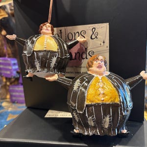May include: Two ornaments depicting a person with outstretched arms. They feature a brown and white striped suit, a yellow shirt, and a brown hairstyle. The ornaments are suspended by a string.