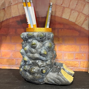 May include: A unique, textured pencil holder in the shape of a creature's foot. The holder is gray with yellow accents and holds several pencils. The pencils have yellow, white, and purple stripes. One pencil reads "EXCELLENT".