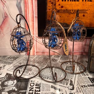 May include: Three blue fairy figurines are each enclosed within a small, round, brown wire cage that hangs from a matching metal stand. Each cage features a hook at the top and a circular base. A small tan tag is attached to one of the wire cages.