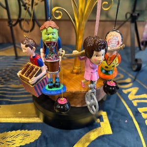 Puede incluir: Una colección de adornos navideños caprichosos. Los adornos presentan figuras en miniatura, incluyendo un ciclista en monociclo, una figura con un tambor y una figura con un ábaco. Cada adorno cuelga de un árbol dorado.