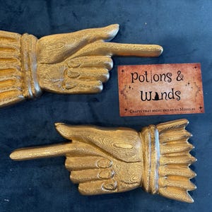 May include: Two gold-colored, hand-shaped wall decorations, each pointing to the right. The hands have detailed textures and are mounted on a dark blue surface. A small card with the text "Potions & Wands" is also present.