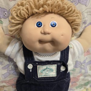May include: A doll with light tan skin, bright blue eyes, and curly blonde yarn hair. The doll wears a white short-sleeved shirt with a decorative stitch at the neckline and dark blue denim overalls featuring a Cabbage Patch Kids label.