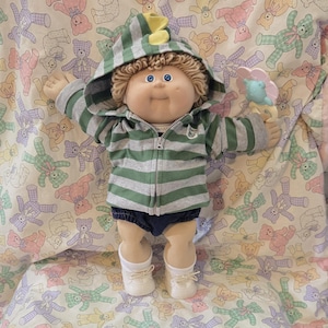 May include: A Cabbage Patch Kids doll with yarn hair and blue eyes, wearing a green and gray striped zip-up hoodie with a yellow dinosaur crest on the hood, dark blue shorts, and white shoes, holding a small pink and blue flower-shaped rattle.