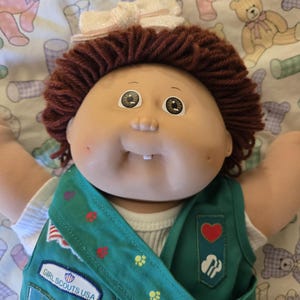May include: A Cabbage Patch Kids doll with brown yarn hair and a white hair bow. The doll wears a white shirt under a green Girl Scouts USA vest featuring a red heart patch, a trefoil logo, and a rectangular patch with the text Girl Scouts USA.