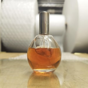 May include: A spherical glass perfume bottle filled with an amber-colored liquid. The bottle features a smooth, rounded body and a cylindrical metallic silver cap at the top. A clear plastic spray tube is visible inside the glass container.