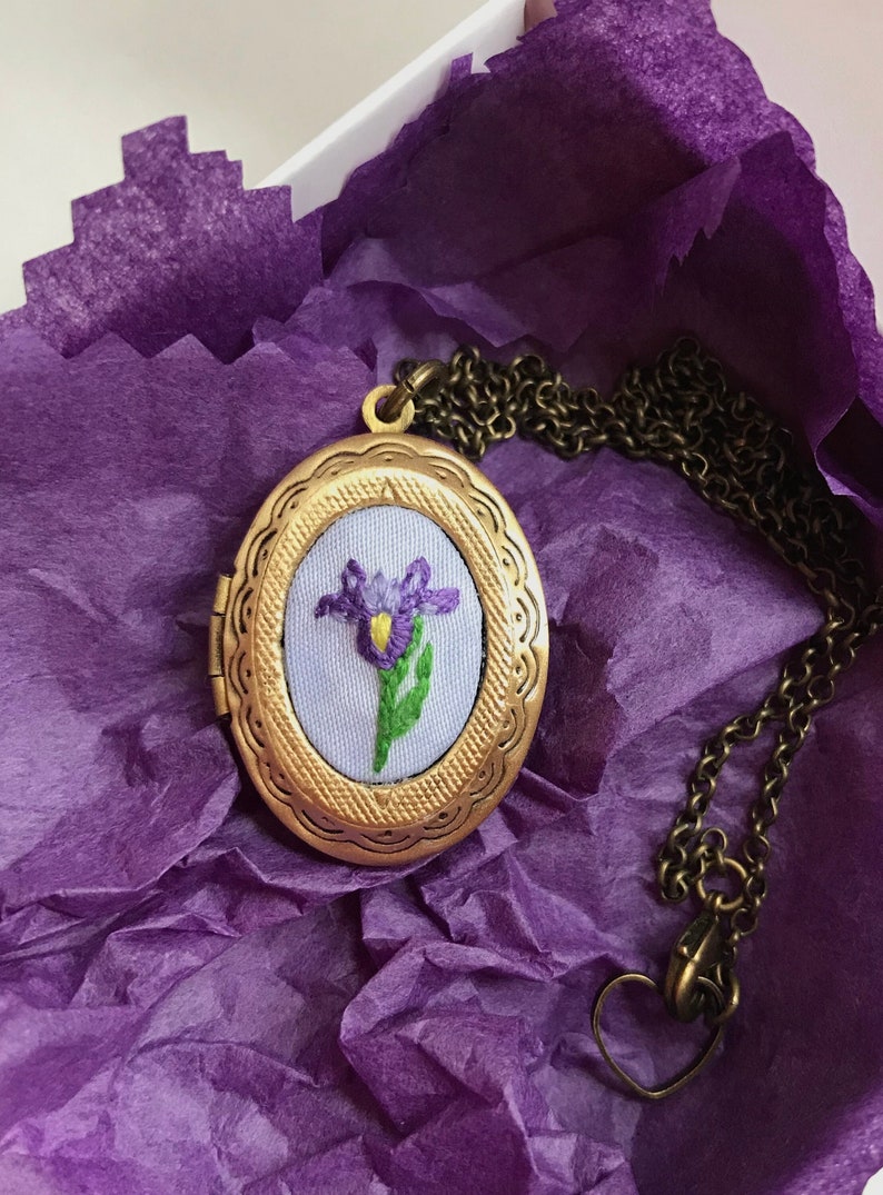 May include: A gold-colored oval locket necklace with a chain and a heart-shaped clasp. The locket features an embroidered purple iris flower on a light blue background, set against purple tissue paper.