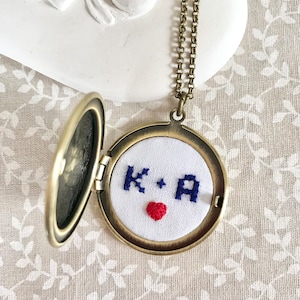 Peut inclure: Un collier avec un médaillon en or, l'intérieur en tissu blanc brodé d'un cœur rouge et des initiales "K + A".