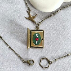 May include: A brass necklace with a locket pendant featuring a green embroidered heart with the letters "S" and "T" and a red heart. The locket is attached to a brass chain with a small bird charm. The necklace also includes a brass lobster clasp and a hexagonal jump ring.