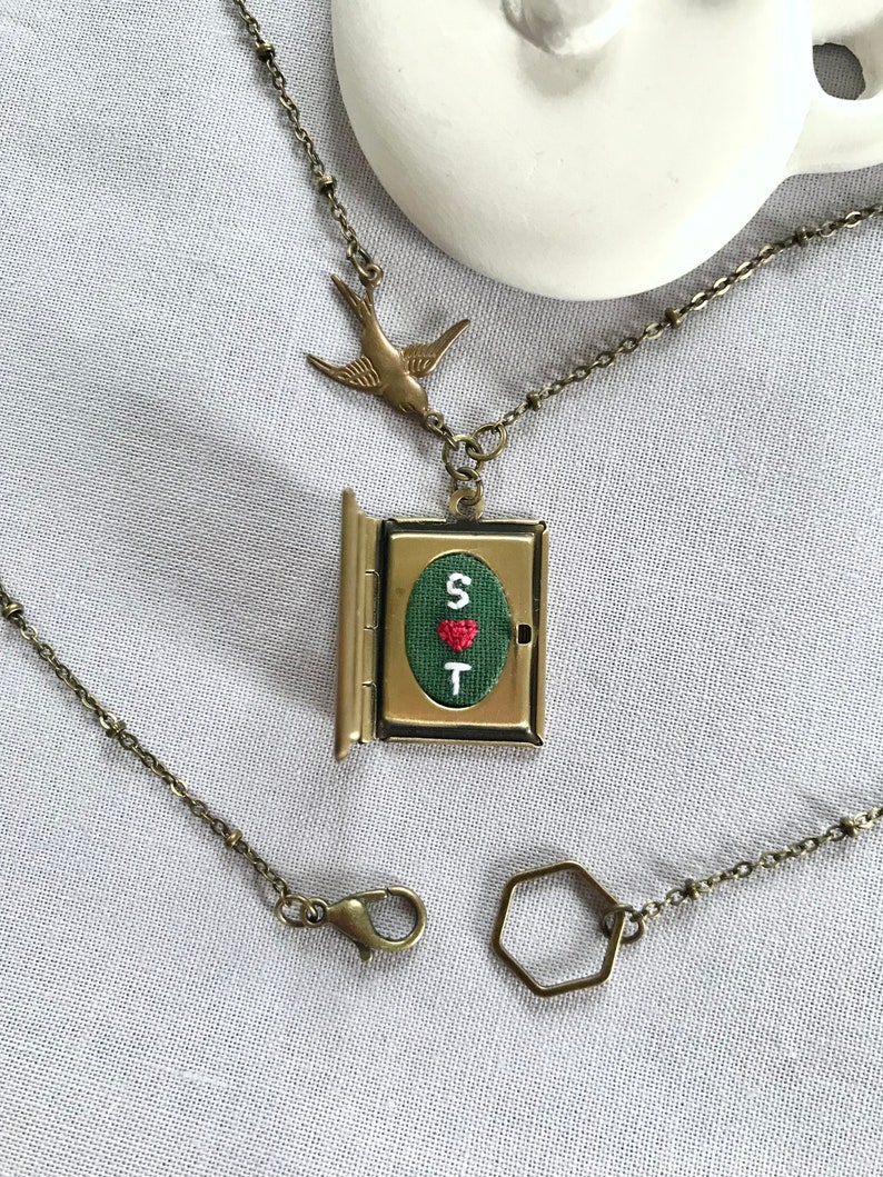 May include: A brass necklace with a locket pendant featuring a green embroidered heart with the letters "S" and "T" and a red heart. The locket is attached to a brass chain with a small bird charm. The necklace also includes a brass lobster clasp and a hexagonal jump ring.