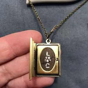 May include: A gold-toned locket necklace with a small book-shaped pendant. The locket is open, revealing a brown fabric interior with the word "LOVE" embroidered in white thread.