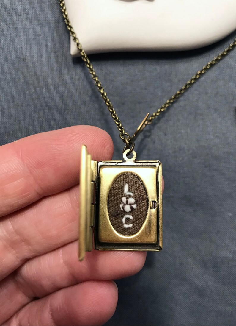 May include: A gold-toned locket necklace with a small book-shaped pendant. The locket is open, revealing a brown fabric interior with the word "LOVE" embroidered in white thread.