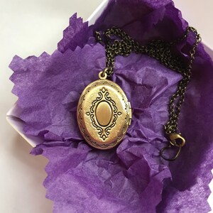 May include: A bronze-colored oval locket necklace with an ornate design, resting on purple tissue paper inside a white box. The chain is dark bronze and has a clasp. The locket is closed and has a decorative border.