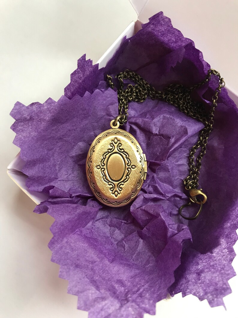 May include: A bronze-colored oval locket necklace with an ornate design, resting on purple tissue paper inside a white box. The chain is dark bronze and has a clasp. The locket is closed and has a decorative border.