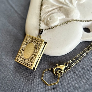 May include: A gold-toned locket necklace with a detailed ornate design. The locket is shaped like a book and has a hinged opening. The necklace has a lobster clasp and a hexagonal extender chain.