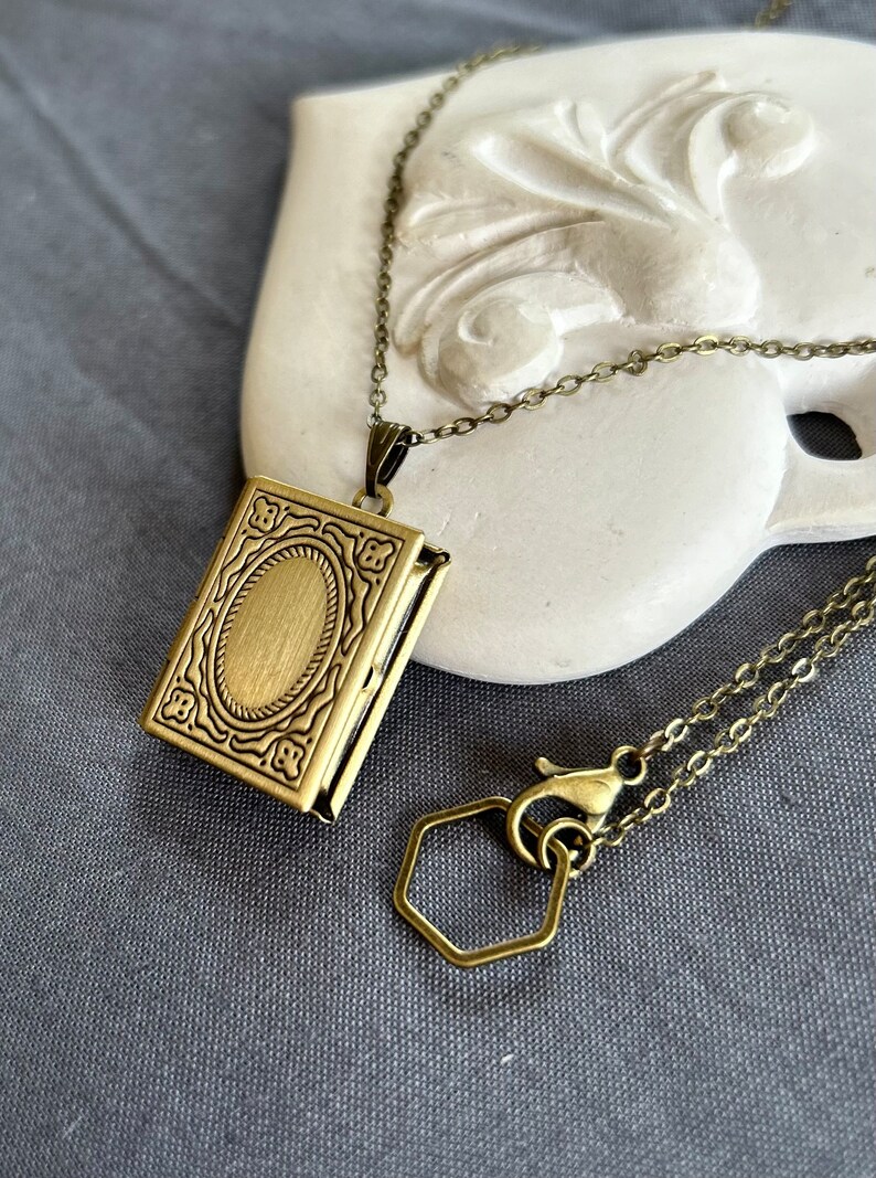 May include: A gold-toned locket necklace with a detailed ornate design. The locket is shaped like a book and has a hinged opening. The necklace has a lobster clasp and a hexagonal extender chain.