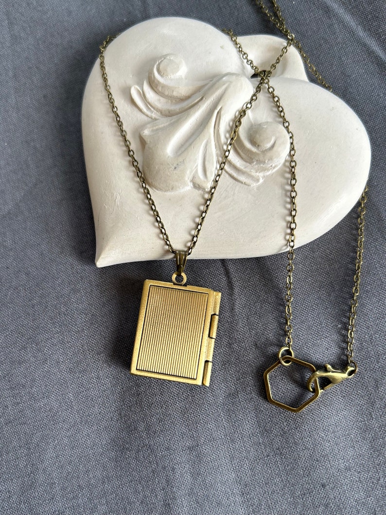 May include: A gold-toned locket necklace with a book-shaped pendant. The necklace has a delicate chain and a hexagonal clasp.