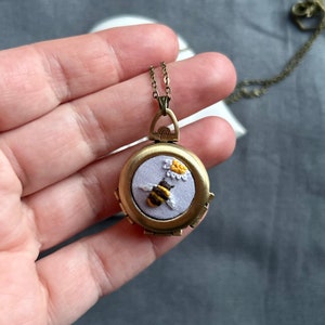 May include: A gold locket necklace with a purple fabric interior featuring an embroidered bee and flower design.