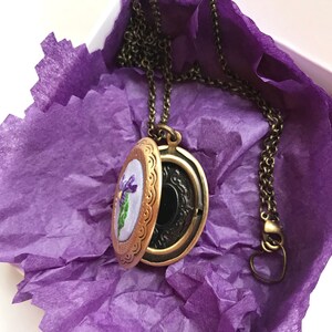May include: An open, antique-style locket necklace with a bronze finish, revealing a floral embroidery. The locket rests on purple tissue paper inside a white box. The chain and clasp are also bronze-toned, creating a vintage aesthetic.