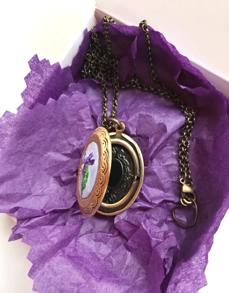 May include: An open, antique-style locket necklace with a bronze finish, revealing a floral embroidery. The locket rests on purple tissue paper inside a white box. The chain and clasp are also bronze-toned, creating a vintage aesthetic.