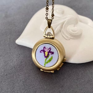 May include: A gold locket necklace with a purple and pink embroidered flower on a white background.