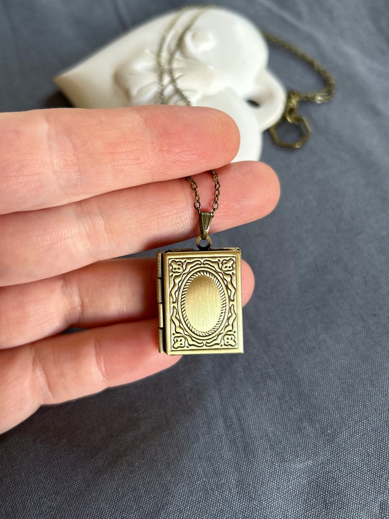 May include: A gold-toned locket necklace with an ornate design. The locket is shaped like a small book and has a hinged opening.