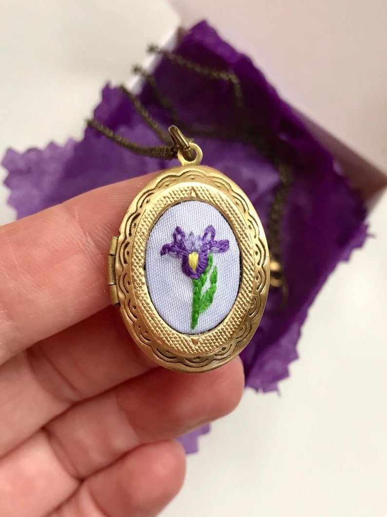 May include: Oval, antique gold-colored locket necklace with an embroidered purple iris flower on a light blue background. The locket is open, revealing the detailed floral design. The necklace chain is visible.