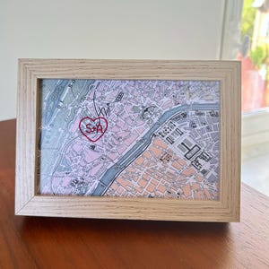 Könnte beinhalten: Ein rechteckiger Rahmen aus hellem Holz mit einer gedruckten Karte von Paris, Frankreich. Die Karte zeigt ein handgesticktes rotes Herz mit den eingestickten Buchstaben S und A, das über einem bestimmten Stadtgebiet mit Straßen und einem Fluss liegt.