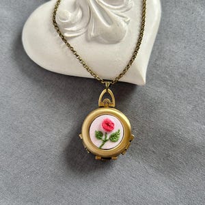 Peut inclure: Un collier avec un médaillon doré orné d'une rose rose brodée sur un fond rose clair. Le médaillon est suspendu à une chaîne fine et repose sur un ornement blanc en forme de cœur.