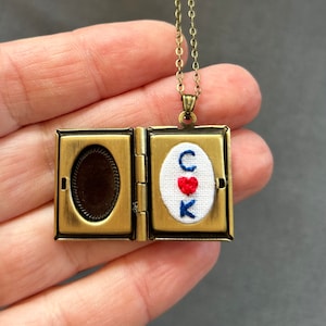 May include: A gold locket necklace with a small embroidered heart and the letters "C K" inside. The locket is open to reveal a blank space inside.