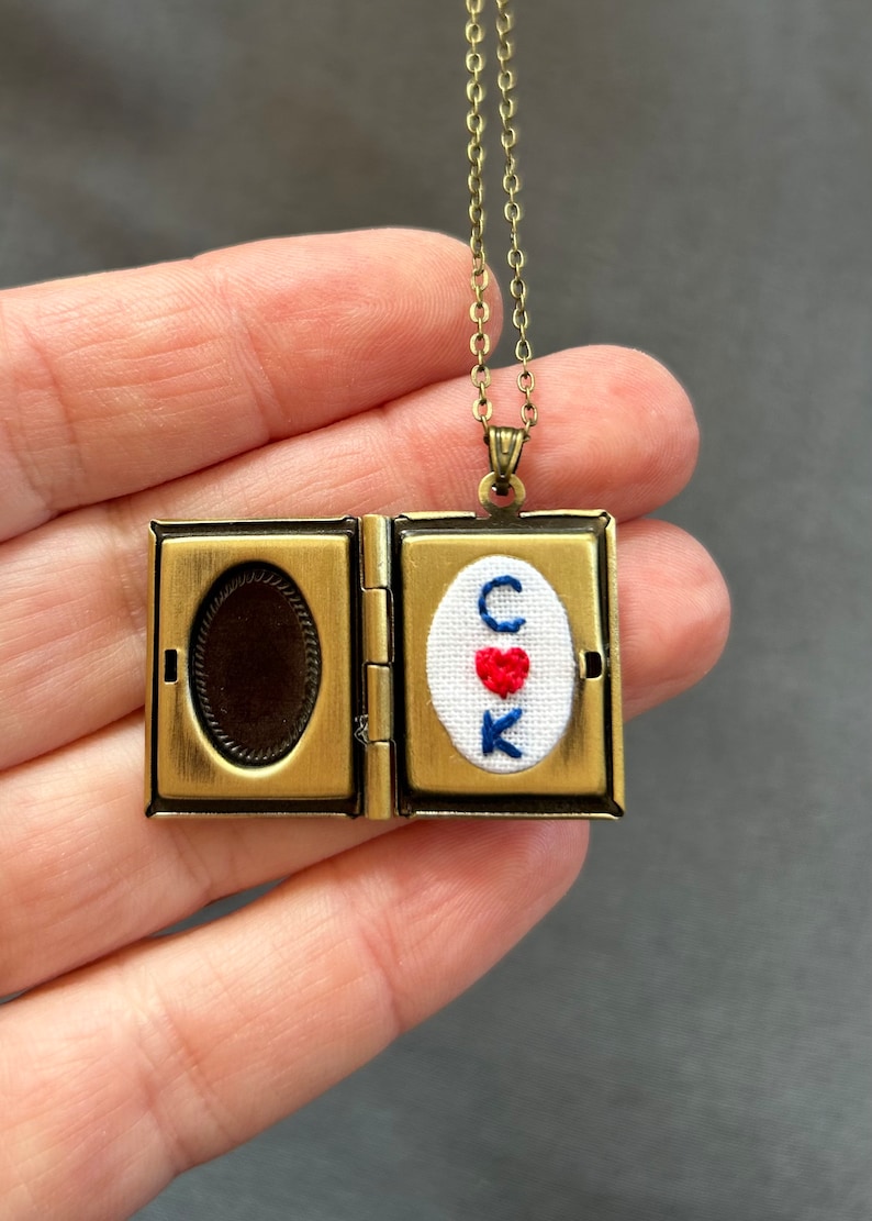May include: A gold locket necklace with a small embroidered heart and the letters "C K" inside. The locket is open to reveal a blank space inside.