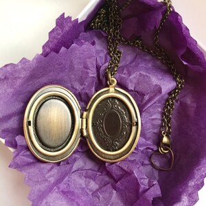 May include: An open, antique-style locket necklace with a bronze finish, resting on purple tissue paper inside a white box. The locket is oval-shaped with an ornate design on the inside. The chain has a heart-shaped clasp.