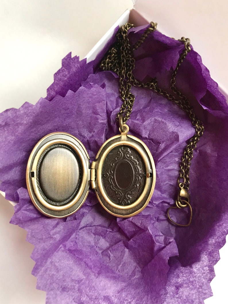 May include: An open, antique-style locket necklace with a bronze finish, resting on purple tissue paper inside a white box. The locket is oval-shaped with an ornate design on the inside. The chain has a heart-shaped clasp.