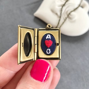 May include: A gold locket necklace with a small, blue fabric heart inside. The heart has a red embroidered heart and the letters "A of O" embroidered in white.
