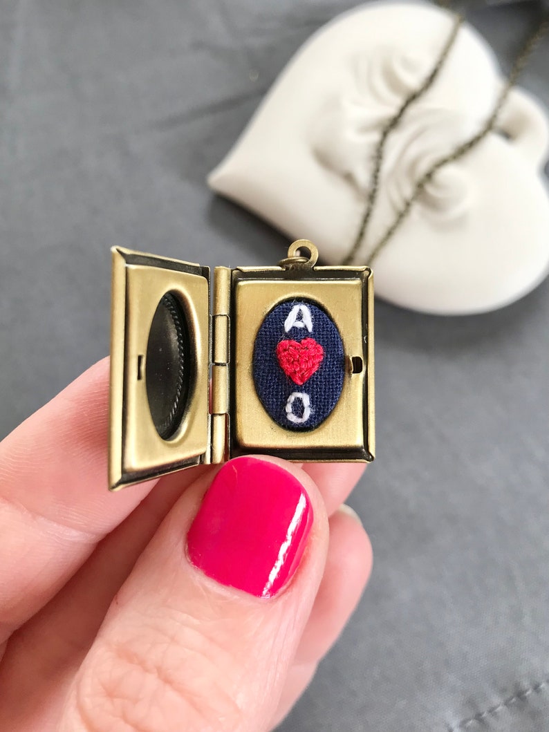 May include: A gold locket necklace with a small, blue fabric heart inside. The heart has a red embroidered heart and the letters "A of O" embroidered in white.