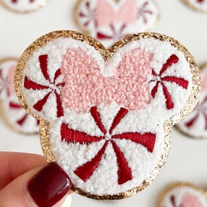 May include: A white and red peppermint candy-themed patch in the shape of a mouse head with a pink bow. The patch has a gold glitter border and is made of embroidered fabric.
