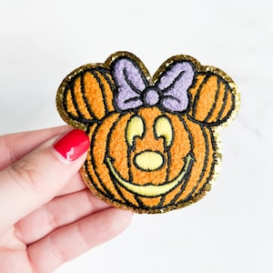 May include: Orange and yellow felt pumpkin with a purple bow and black eyes. The pumpkin has a stitched-on smile. The pumpkin is surrounded by a gold glitter trim.