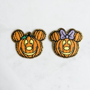 以下が含まれることがあります： ミッキーマウスとミニーマウスがカボチャになった、ハロウィンをテーマにした刺繍パッチ2つ。パッチはオレンジ色で、金色のグリッターのアクセントが施され、緑色の茎と紫色のリボンが縫い付けられています。