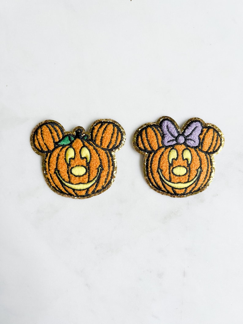 以下が含まれることがあります： ミッキーマウスとミニーマウスがカボチャになった、ハロウィンをテーマにした刺繍パッチ2つ。パッチはオレンジ色で、金色のグリッターのアクセントが施され、緑色の茎と紫色のリボンが縫い付けられています。