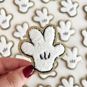 May include: A white fuzzy fabric Mickey Mouse glove patch with a gold glitter outline. The glove has black stitching for the fingers and a black outline around the wrist.