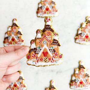 May include: Embroidered gingerbread house patches with gold trim, featuring candy cane accents, red hearts, and white frosting details. The patches are in the shape of a castle, with a Mickey Mouse head design.