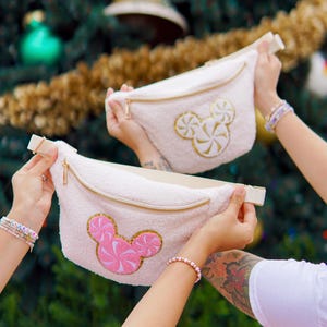 May include: Two white fuzzy fanny packs with a Mickey Mouse head design featuring a pink and white peppermint swirl. The fanny packs have gold zippers.