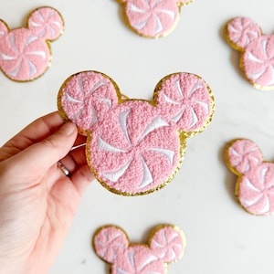 May include: Pink and white embroidered patches in the shape of a mouse head with a gold glitter border. The patches have a candy swirl design. Several patches are scattered on a white surface.
