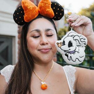 May include: A woman wearing a black and orange sequined bow headband with black ears and gold polka dots. She is holding a white plush Zero keyring with a gold ring. A gold necklace with a pumpkin pendant is also visible.