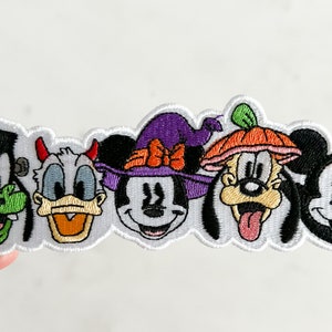 May include: An embroidered patch featuring five Disney characters dressed in Halloween costumes. The characters are Mickey Mouse, Minnie Mouse, Donald Duck, Goofy, and Pluto. Mickey Mouse is wearing a black and white witch's hat, Minnie Mouse is wearing a purple witch's hat with a red bow, Donald Duck is wearing a red devil's hat, Goofy is wearing an orange pumpkin hat, and Pluto is wearing a green leaf hat.