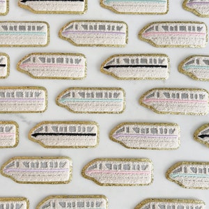 May include: A collection of 18 gold-trimmed embroidered patches featuring a white monorail train with different colored stripes. The patches are arranged in a grid pattern on a white background.