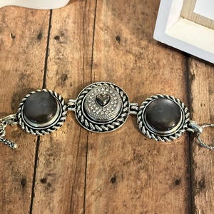 May include: A silver-toned bracelet with three circular decorative elements. The outer two elements have a dark gray stone center, while the center element features a heart-shaped design surrounded by clear crystals. The bracelet has a toggle clasp.