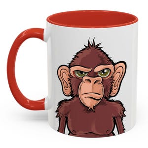 May include: A white ceramic coffee mug with a red rim and handle. The mug features a cartoon illustration of a brown monkey with a serious expression.