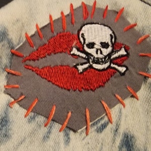 Könnte beinhalten: Ein grauer Stoff-Patch mit roten Nähten an den Rändern. Der Patch zeigt einen weißen Totenkopf mit gekreuzten Knochen und einem roten gestickten Lippen-Design.