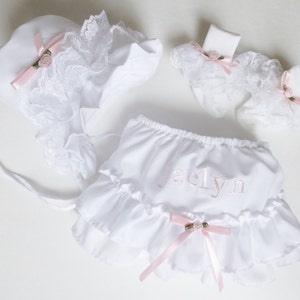Peut inclure: Ensemble bébé blanc composé d'une jupe à volants, d'un chapeau et de chaussettes. La jupe est ornée d'un nœud rose et du nom "Jaelyn" brodé. Le chapeau et les chaussettes sont en dentelle rose et ont des nœuds.